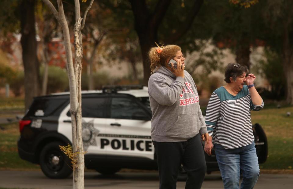 A woman wipes away tears as another speaks on a cellphone. In the background is a police car.