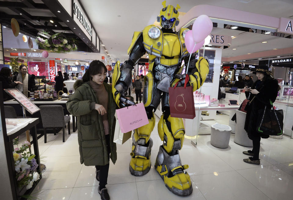 In this Thursday, Feb. 14, 2019, photo, a Transformer's Bumblebee mascot helps a woman carries paper bags at a shopping mall in Handan in north China's Hebei province. U.S. and Chinese envoys are holding a second day of trade talks after the top economic adviser to President Donald Trump said he has yet to decide whether to go ahead with a March 2 tariff increase on imports from China. (Chinatopix via AP)