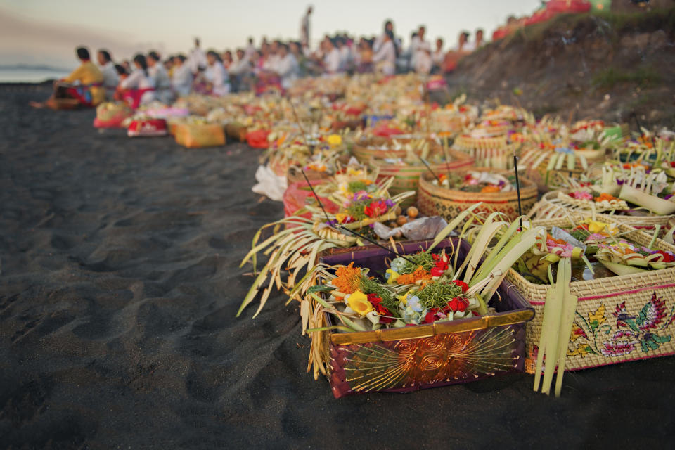 Baskets filled with flowers and offerings are arranged on a beach for a ceremonial event, with a group of people gathered in the background