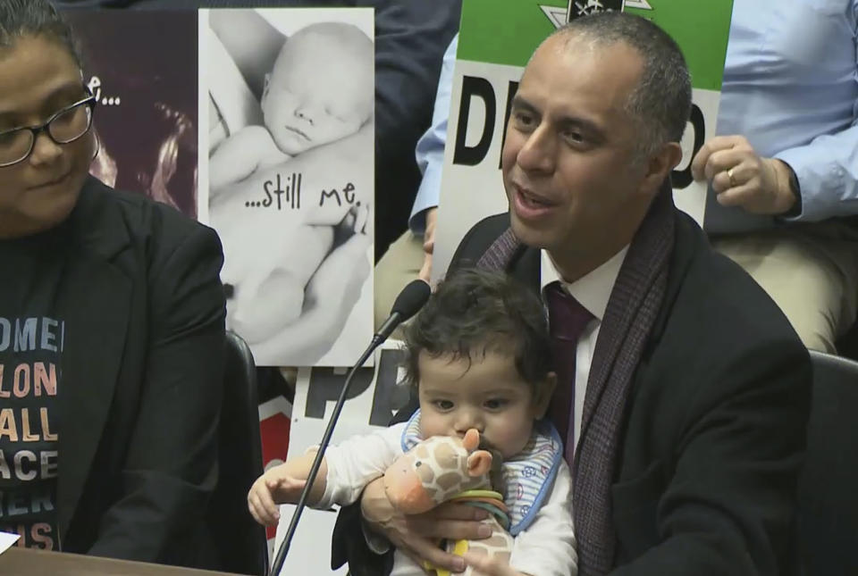 In this Jan. 29, 2019, frame from video from Capitol TV, Mayor Jorge Elorza holds his son Omar as he testifies before the House Judiciary Committee at the Statehouse in Providence, R.I. Elorza's decision to frequently take his son to work with him has brought praise from some who say he's a role model, but also raised questions about whether he has taken it too far. (Capitol TV via AP)