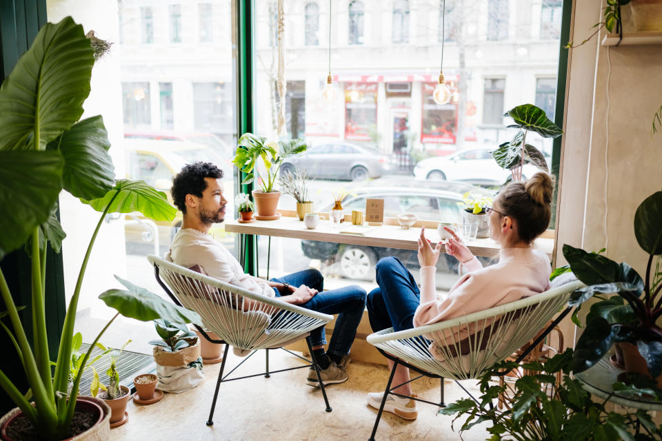 A couple hanging out in a cafÃ©, sitting by a large window with lots of plants around.