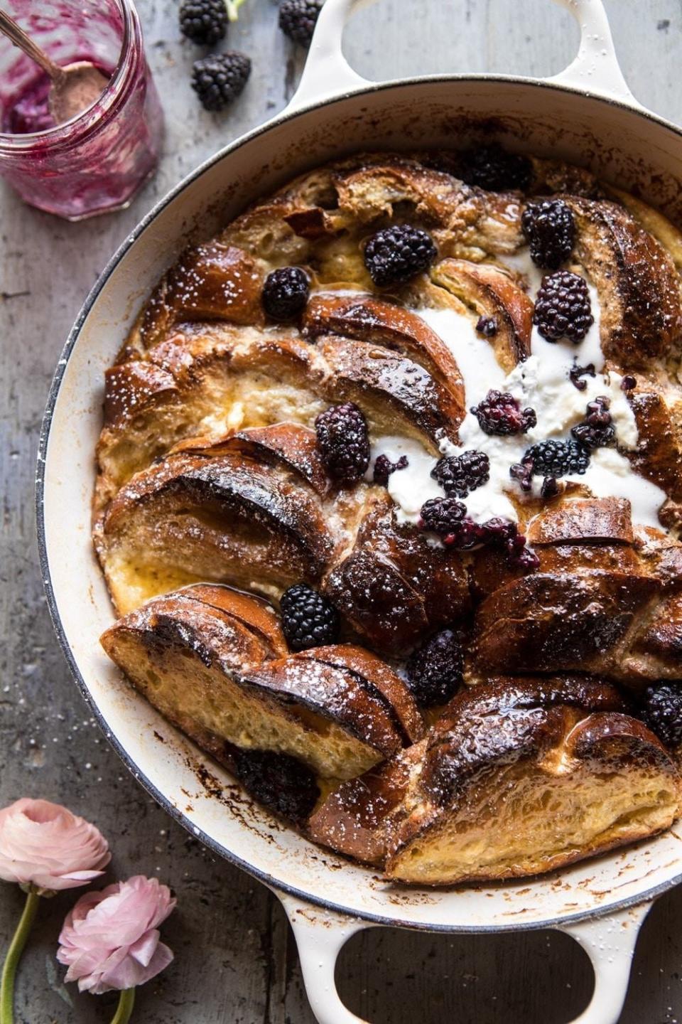 French toast baked with blackberries and cream in a white dish, garnished with powdered sugar and accompanied by a jar of jam and flowers