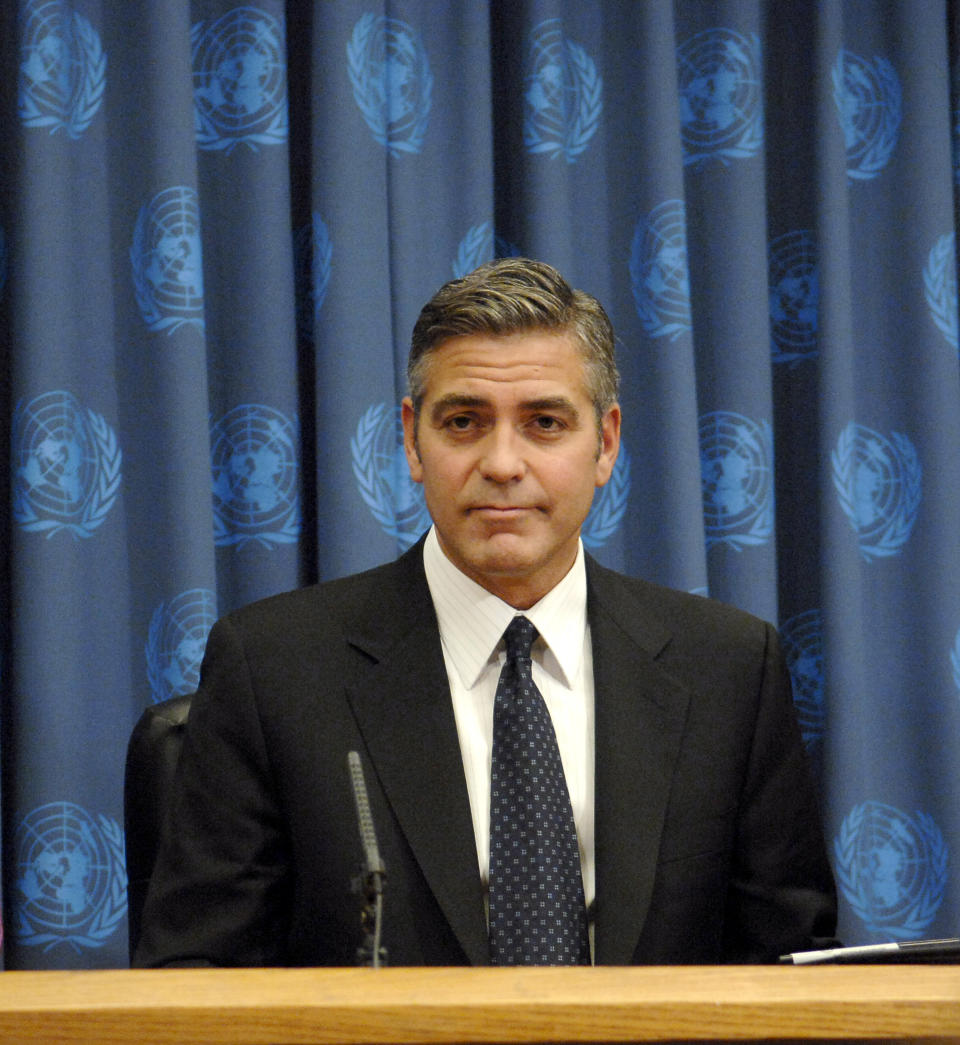 Close-up of George wearing a suit and tie with a curtain behind him