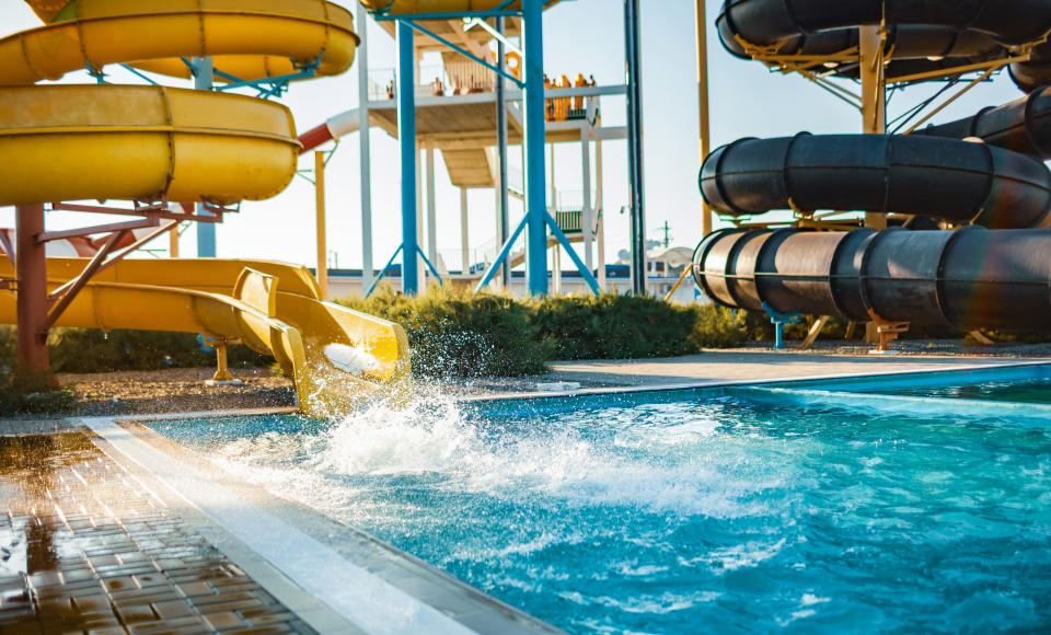 Water park with various slides, including a yellow slide splashing into a pool, and people in the distance watching from a platform. Sunlit, outdoor scene