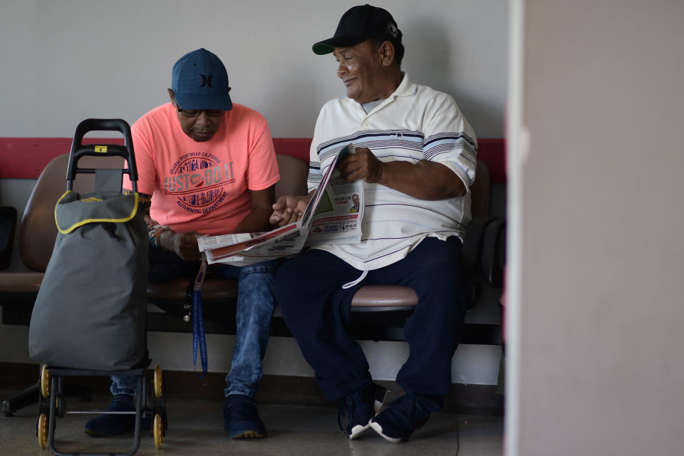 In this Sept. 8, 2018 photo, dialysis patients Elias Salgado, 56, left, and Edwin Alvarado, 59, banter as they read a newspaper while waiting to board a plane in Vieques, that will fly them to the Puerto Rican mainland for treatments. Salgado's doctor said he worries about the effects of such exhausting travel. "Eventually those patients, who already are fragile, will keep worsening," he said, calling a year worth of flights "unacceptable". " (AP Photo/Carlos Giusti)