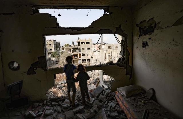 Children standing on rubble in a bedroom look through a shattered wall at ruined buildings.