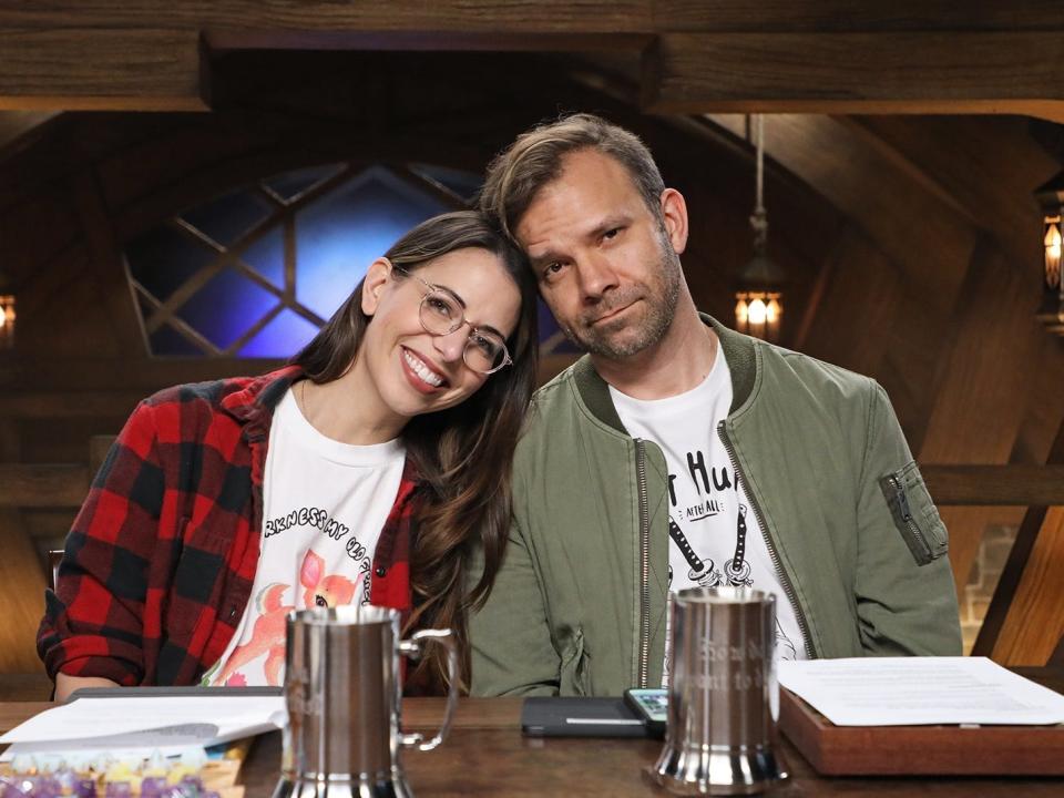 Liam O'Brien and Laura Bailey pose for a photo in Critical Role's studio. Bailey is wearing a T-shirt with a red checkered jacket, and O'Brien is wearing an olive green bomber jacket with a white t-shirt. 