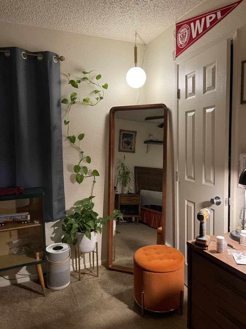 Corner of a bedroom with a standing mirror, air purifiers, and orange ottoman on a carpeted floor.