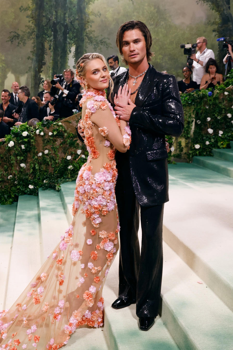Two individuals at an event. Person one in a floral-embellished sheer gown; person two in a sparkling black suit. Photographers in the background