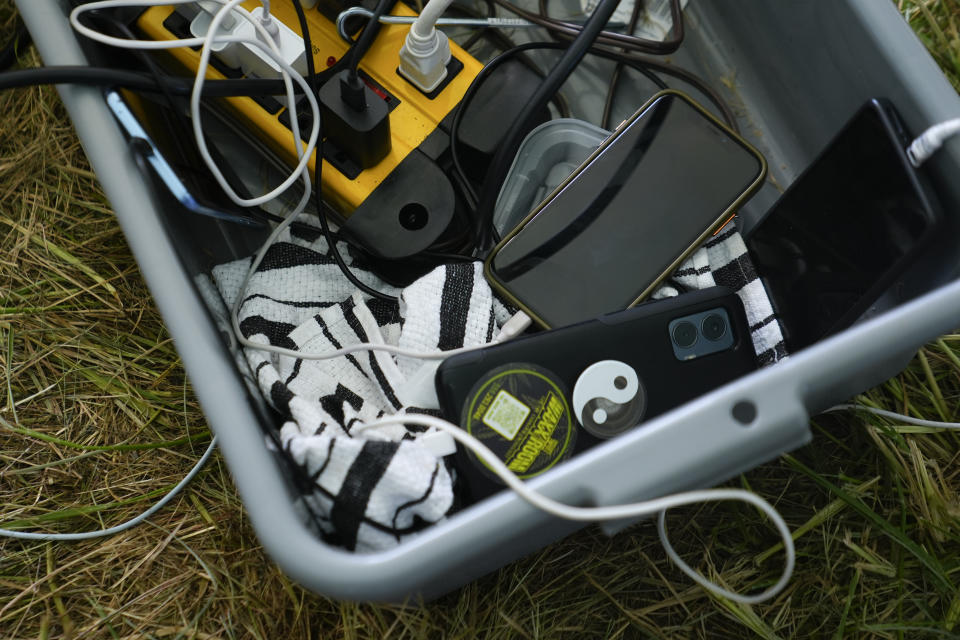 Cellphones sit in a bucket at a makeshift charging station connected to a small generator at an encampment of asylum-seekers next to an unused motel owned by the county, Wednesday, June 5, 2024, in Kent, Washington. The group of about 240 asylum-seekers is asking to use the motel as temporary housing while they look for jobs and longer-term accommodations. (AP Photo/Lindsey Wasson)