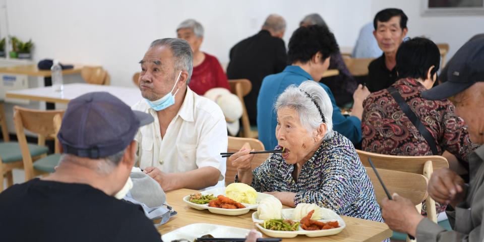 Elderly Chinese people sitting at dining tables and eating plates of food.