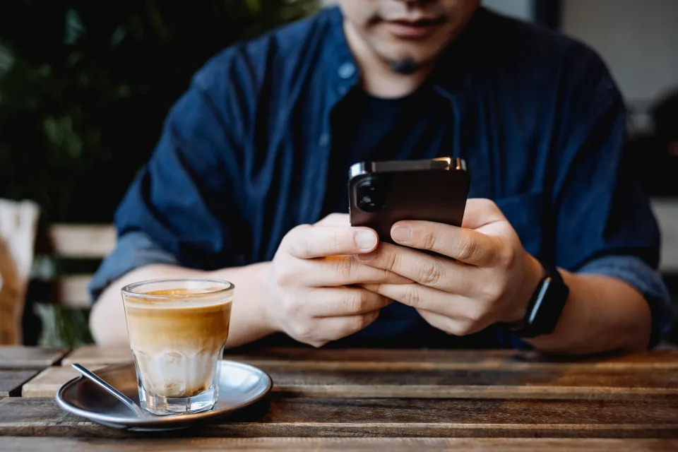 Cropped shot, mid-section of young Asian man sitting at a table in cafe, using smartphone while having a cup of coffee. Lifestyle and technology