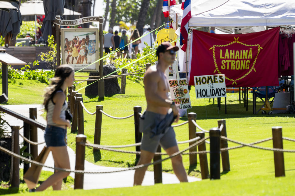 People walk by a tent with a "Lahaina strong" flag, Wednesday, Dec. 6, 2023, at Whalers Village on Kaanapali Beach in Lahaina, Hawaii. Residents and survivors still dealing with the aftermath of the August wildfires in Lahaina have mixed feelings as tourists begin to return to the west side of Maui, staying in hotels still housing some displaced residents. A group of survivors is camping on the resort beach to protest and raise awareness for better long-term housing options for those displaced. (AP Photo/Lindsey Wasson)