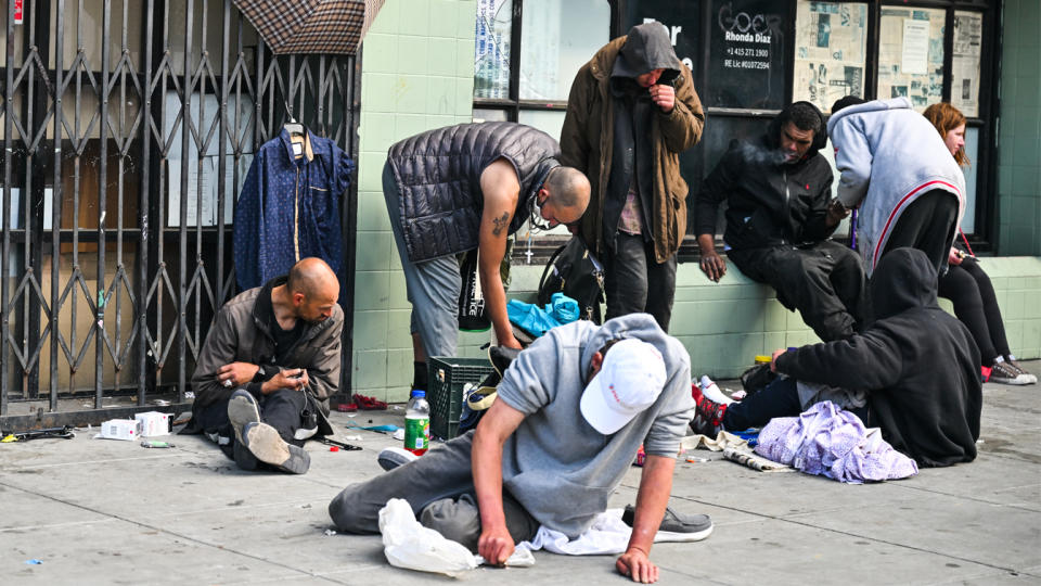 Eight people, apparently destitute, sit on the sidewalk beside the metal grille of a shuttered store.