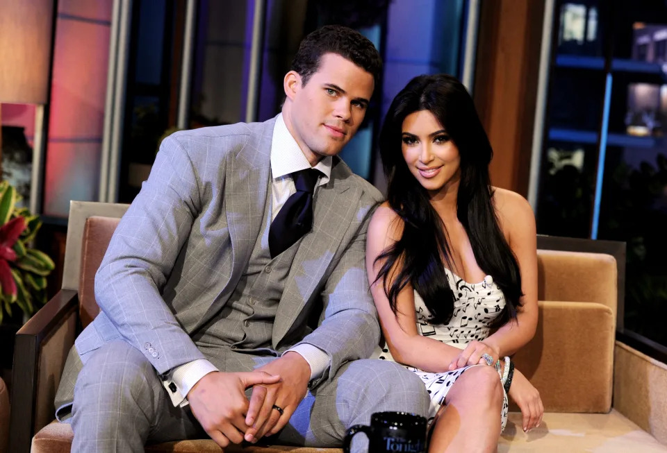 Kris Humphries and Kim Kardashian sit together on a talk show set. Kris wears a grey suit and Kim wears a patterned dress