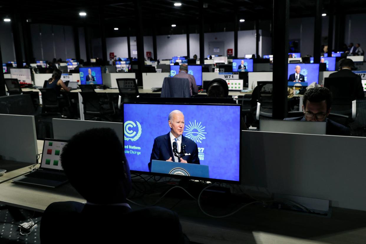Journalists, seen from behind, seated in front of large computer screens showing President Biden.