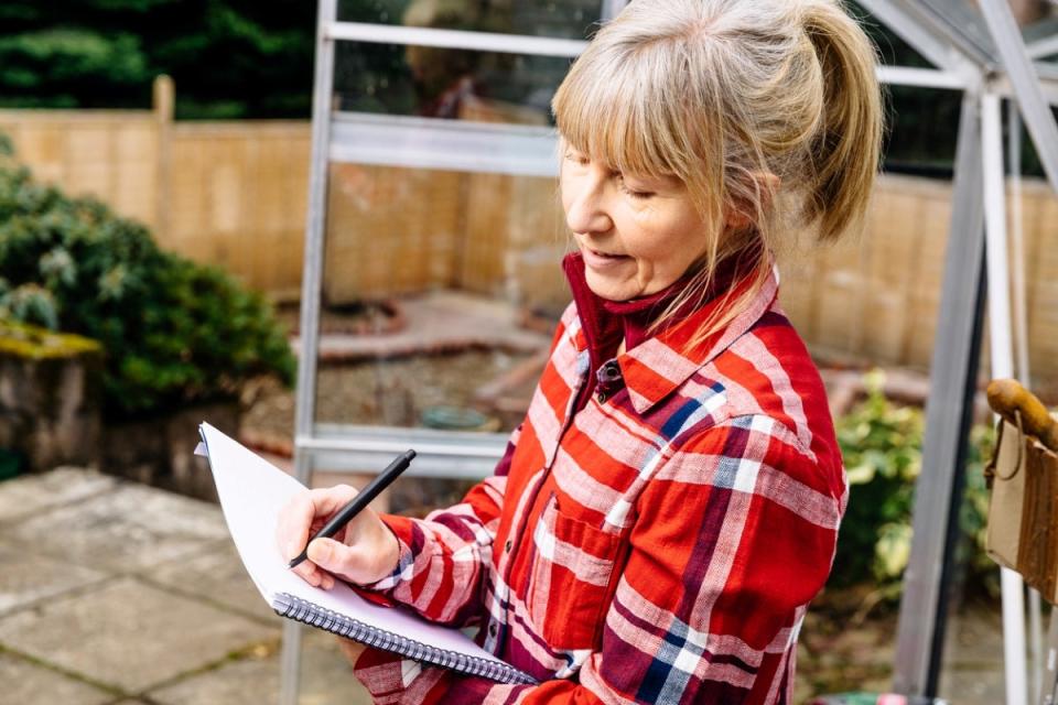 Woman wearing a red and white shirt writing garden plans in notebook outdoors.