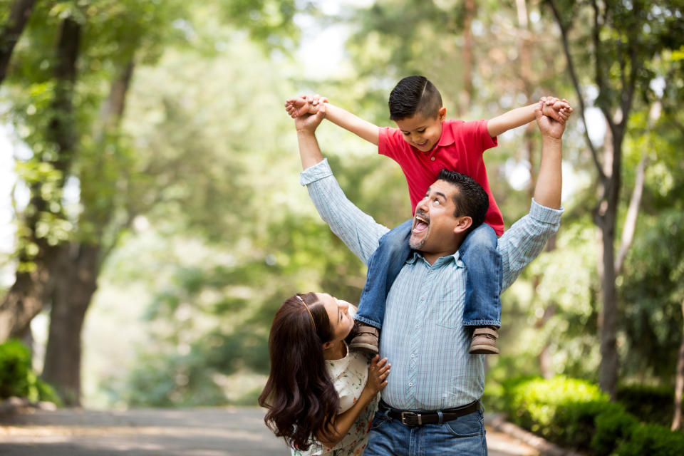 A latin man standing next to wife, carrying his son on his shoulders and smiling at each other in a horizontal medium shot outdoors.