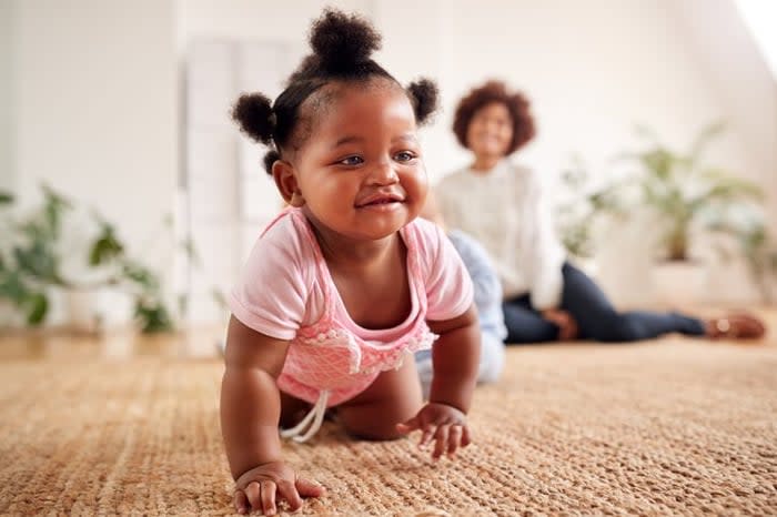 A smiling baby crawling across the floor while the mom sits behind her.
