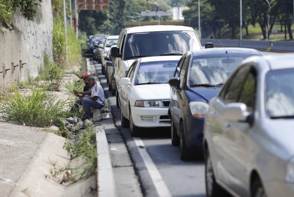 Men sit on a curb alongside vehicles lined up to enter a gas station during a nation-side fuel crunch, in Caracas, Venezuela, Wednesday, Sept 30, 2020. (AP Photo/Ariana Cubillos)