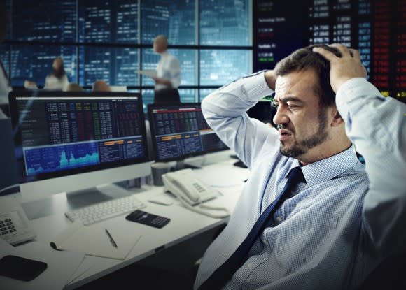 A frustrated man sitting at a desk in front of a computer grasps his head while looking at big losses on his screen.