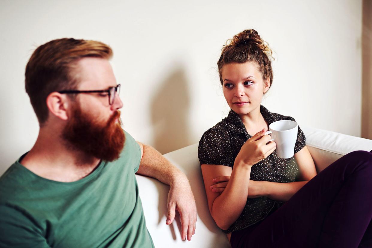 Young man and woman sitting on couch, man looking down while talking towards woman, attentive woman is looking at his eyes, while holding a cup of tea, her arms are crossed, a white wall in the background with a sliver of wooden door frame on the right