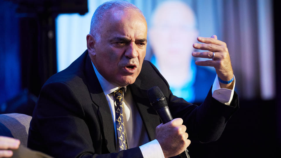 Garry Kasparov, seated, holds a microphone with his right hand and gestures with his left.