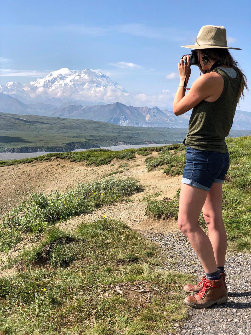 Emily, wearing a hat, a green tank top, shorts, and hiking boots, takes a photo with grass and mountains in the background. 