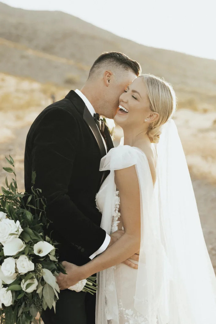 A bride laughs and holds a bouquet as her groom kisses her cheek.