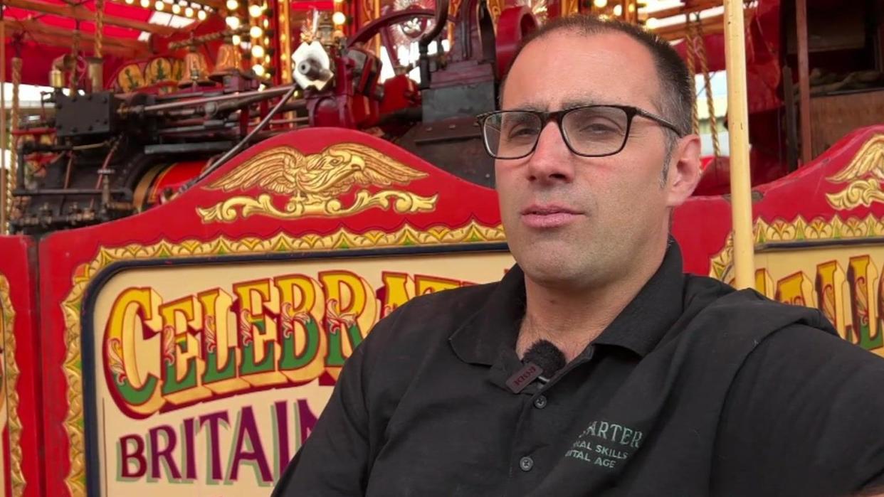 Joby Carter wearing a black top and glasses, standing in front of a colourful painted fairground ride