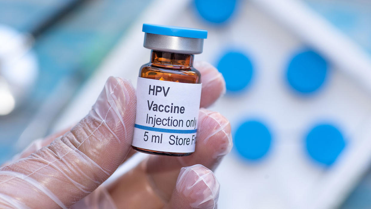 A gloved hand holds a vial of human papillomavirus vaccine. 