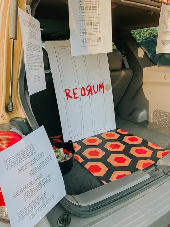 open tailgate revealing large square of paper with the orange and black wallpaper pattern from the shining on it in front of a drawing of a door with redrum in red written on it and hanging around the open tailgate are papers with all work and no play makes jack a dull boy typed on it