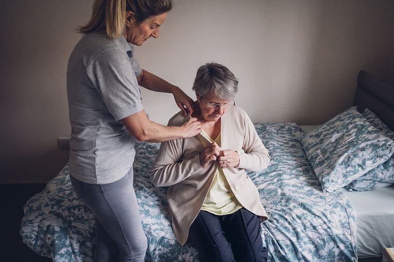 A younger woman is providing care for an elderly lady who is sitting on a bed.