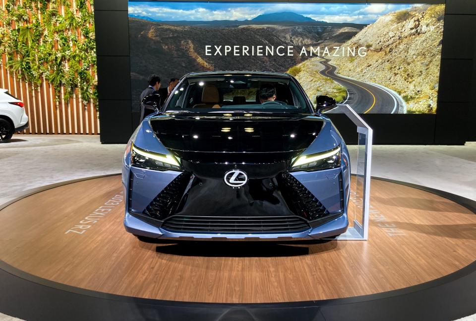 A blue Lexus RZ450e on a wood floor, with a large LED screen in the background reading "experience amazing."