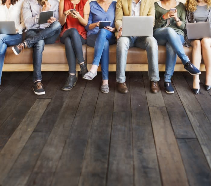 A line of people sitting on a bench using laptops, tablets, and smartphones.