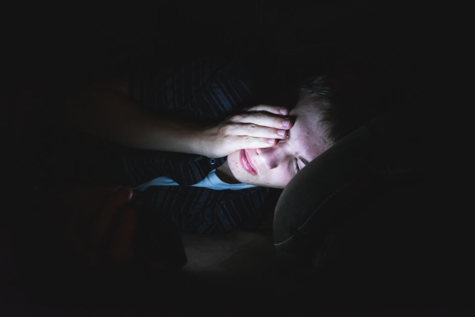 A person lying down in dim light, covering one eye with their hand while looking at a screen