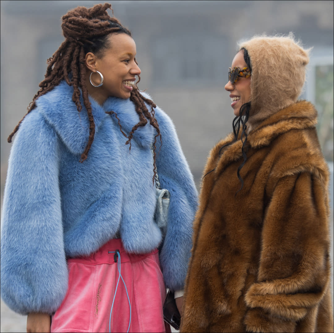  Two women stand side by side, one wearing a blue faux fur coat and another wearing a brown faux fur coat. 