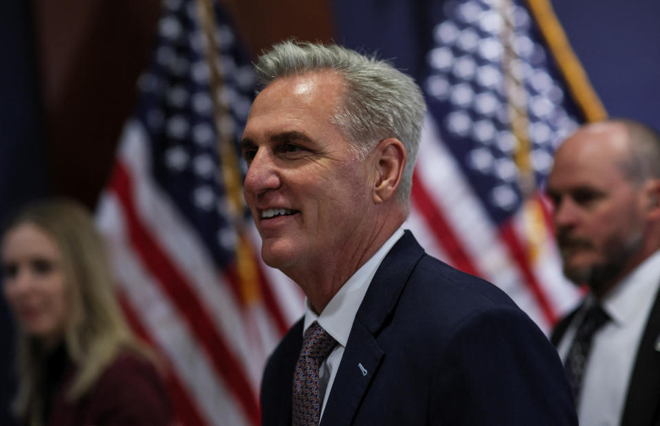 Kevin McCarthy walks forward with enthusiasm, flanked by a woman and a man, with a backdrop of U.S. flags.