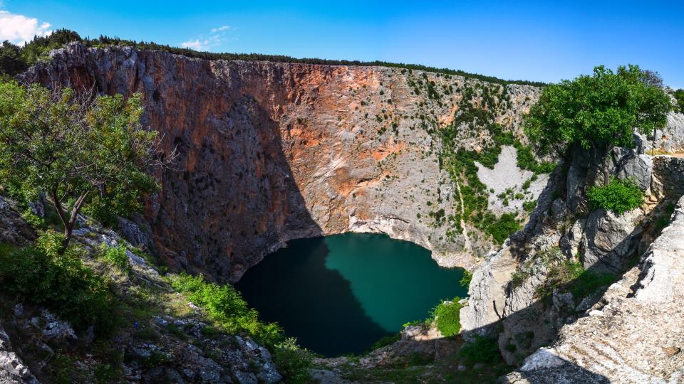 A very large sinkhole with a lake at the bottom with red-colored rocks at the sides