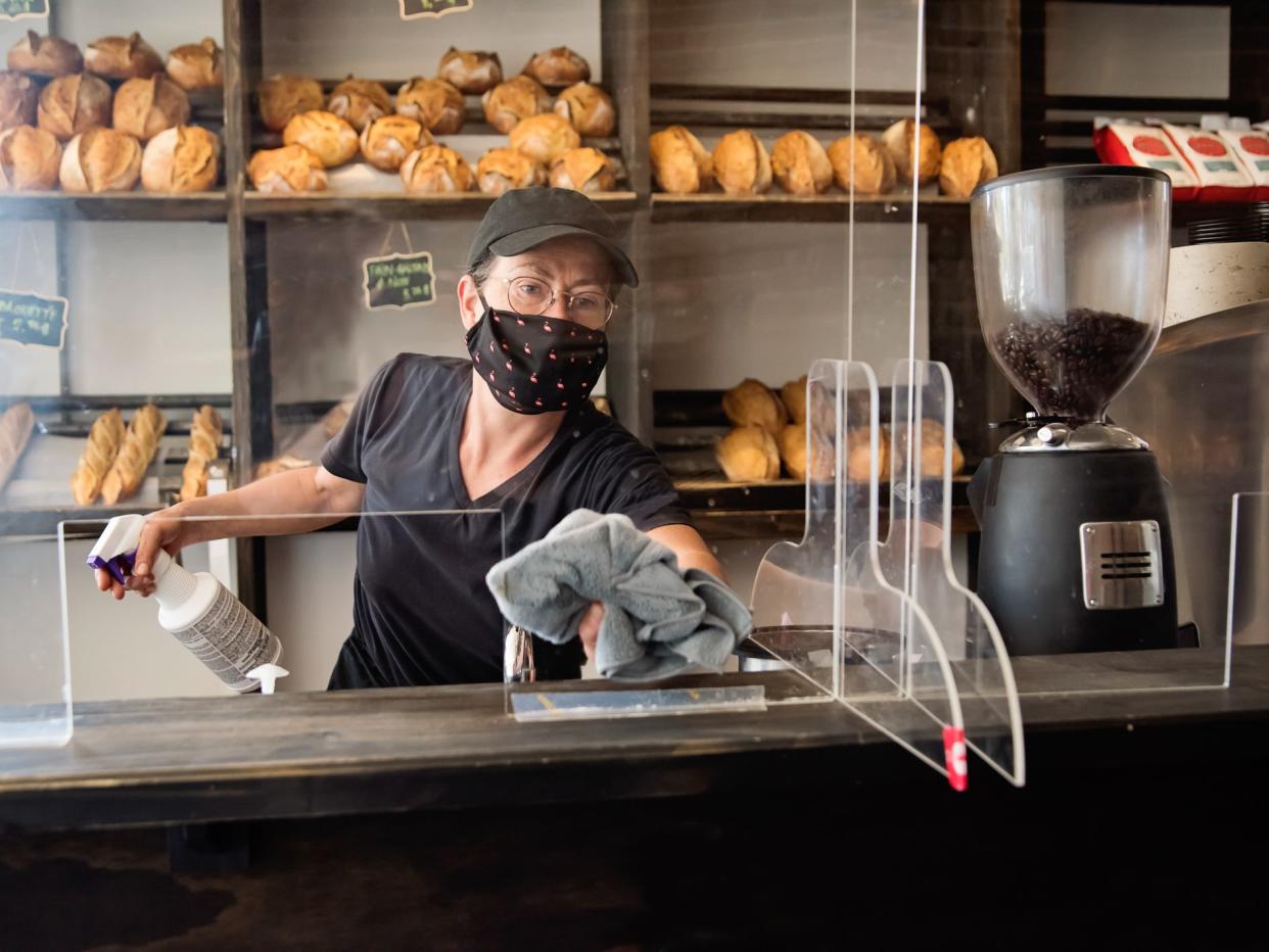 Bakery owner wiping down surfaces wearing mask, following protocol against the propagation of Covid-19. She is dressed in black wiping the protective plexiglass. Horizontal waist up indoors shot with copy space.