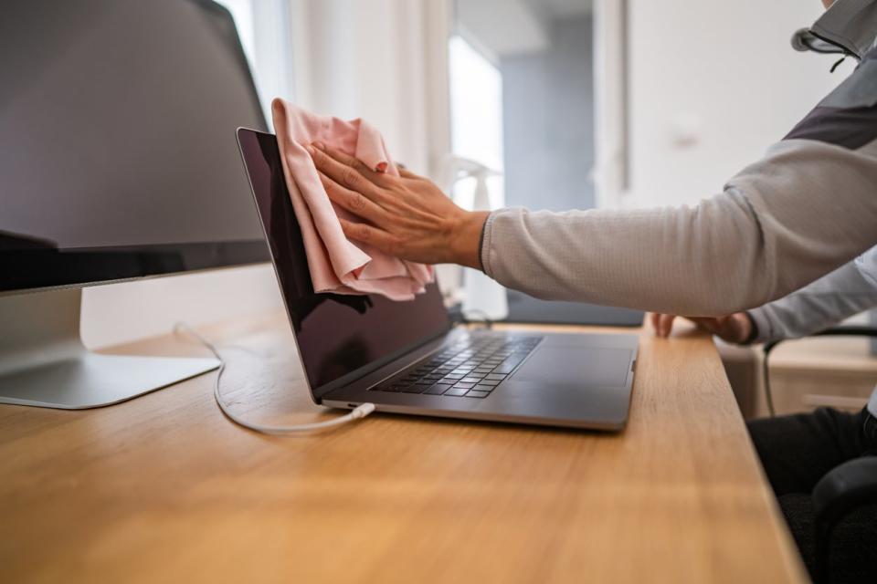 A home office worker wiping off her charging laptop's screen with a pink microfiber cloth.