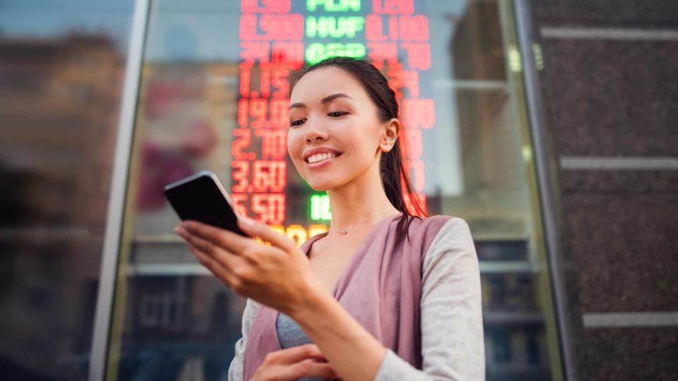 A young beautiful Asian woman using an application in her smart phone to check currency exchange rates in front of an illuminated information board.