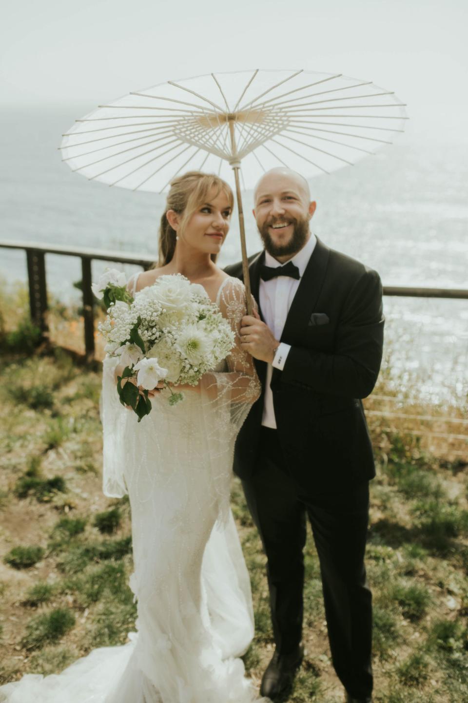 A bride and groom pose in their wedding attire under a parasol.