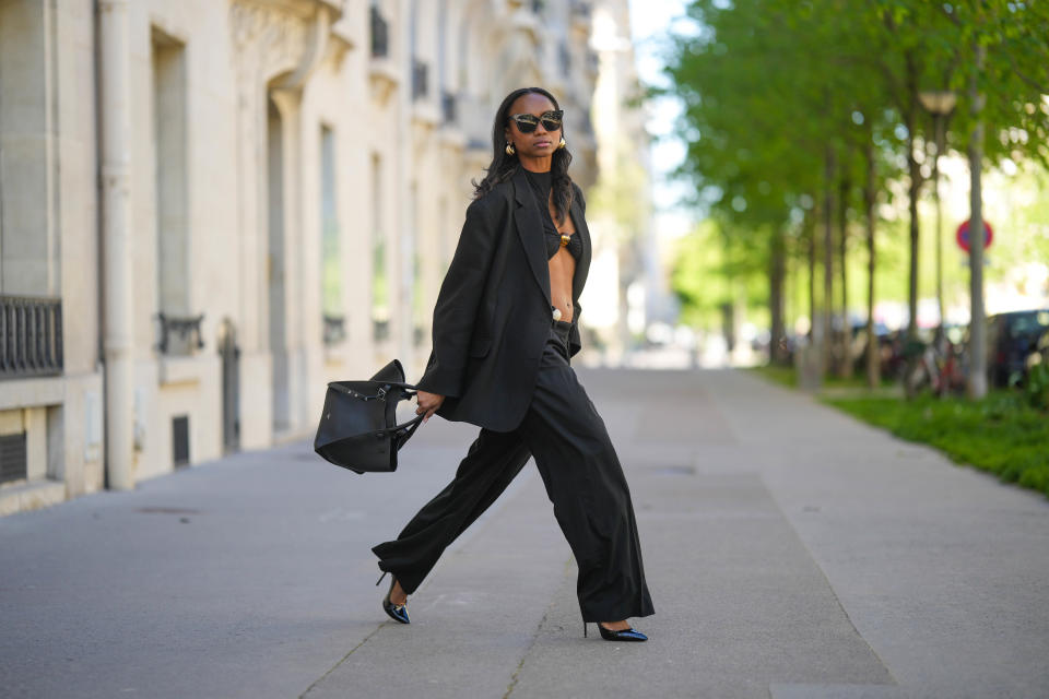 PARIS, FRANCE - APRIL 04: Emilie Joseph wears black sunglasses, gold large earrings, a black cut-out asymmetric shoulder / with a large brown pearl chest bodysuit from Jacquemus, a black oversized blazer jacket The Frankieshop, silver rings, a black grained shiny leather handbag from Celine, black large wide legs pants from The Frankieshop, black shiny leather varnished pointed pumps heels shoes, during a street style fashion photo session, on April 04, 2023 in Paris, France. (Photo by Edward Berthelot/Getty Images)