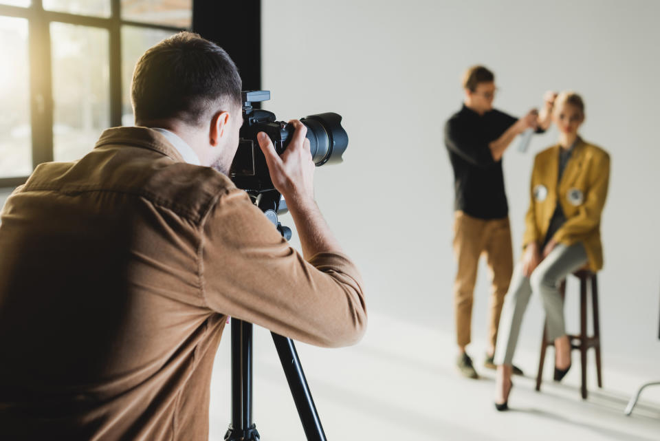Photographer taking a picture of a model seated on a stool with a stylist adjusting her hair