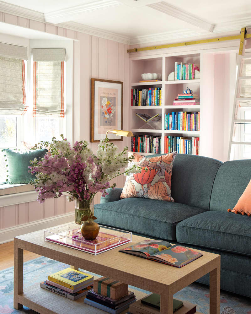 Coffee table with vase of flowers in front of teal loveseat with floral throw pillow in pink library with built-in shelves.