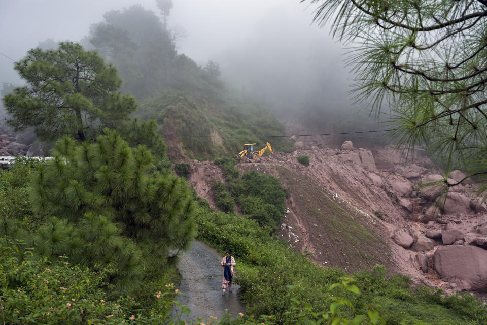 A woman walks down checking her phone as an earth-mover being used to remove the debris after a landslide damaged part of a road near Dharamshala, India, Monday, Aug. 14, 2023. Heavy monsoon rains triggered floods and landslides in India's Himalayan region, leaving several people dead and many others trapped. (AP Photo/Ashwini Bhatia)