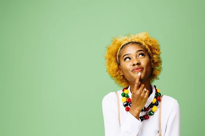 A woman standing in front of a green while while thinking about a decision with her finger resting on her chin.