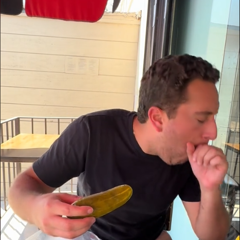 Man holding a pickle and coughing, seated outside at a table with food