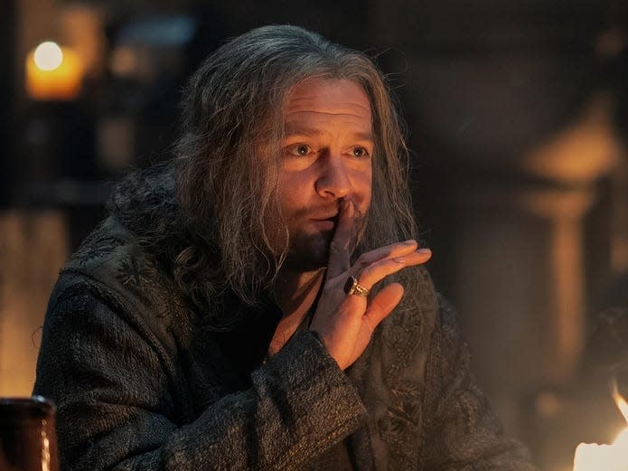 Tom Bennett as Ulf, a middle-aged man with shoulder-length gray hair, placing a finger up to his lips as he sits in a tavern.